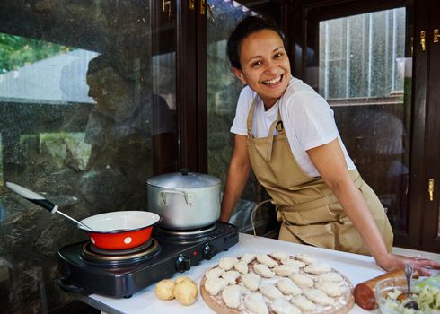 Beautiful dark-haired multi-ethnic woman in beige chef's apron, smiling a cheerful toothy smile, looking at the camera while cooking delicious homemade dumplings in the rustic kitchen. Baking concept