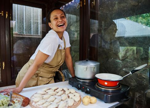 Stunning woman, pleasant young housewife wearing a beige chef's apron, standing by a table with raw dumplings sprinkled with flour and a pot on an electric stove, cooking vareniki in a country kitchen