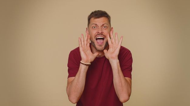Expressive bearded handsome young man holding hands near mouth loudly screaming, shouting, yelling, announces discounts sale Hurry up, welcomes someone hello. Adult stylish guy on beige background