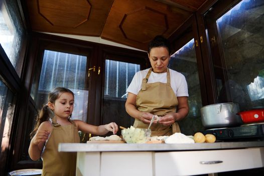 Beautiful child, a little girl in a beige chef's apron, standing next to her mother in the kitchen of a summer cottage, repeating the molding of dumplings, enjoying cooking Ukrainian varenyky together