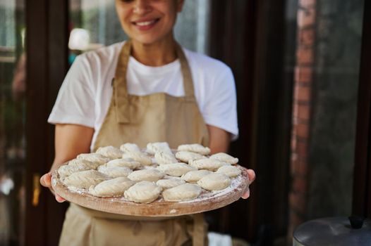 Selective focus on dumplings or vareniki sprinkled with white flour on a wooden board, in the hands of a charming woman housewife in chef's apron, smiling a beautiful toothy smile at the camera