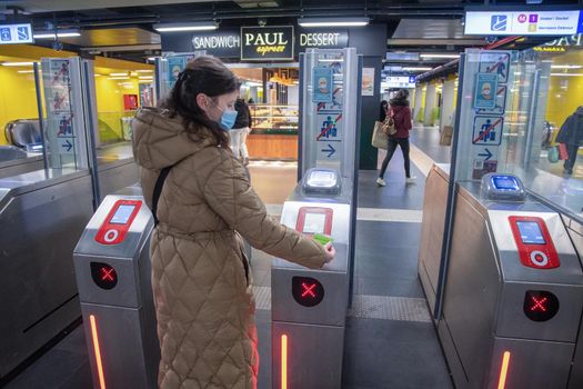Brussels Belgium-01.31.2022 a young woman in a hygienic mask with a warm coat scans a card at the terminal in the subway to exit the platform, sliding doors open in front of her. High quality photo