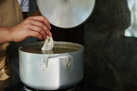 Close-up. Cropped view of a housewife standing by a kitchen table with electric stove, opening the lid of a metal saucepan, dipping raw dumplings into a boiling water, brewing Ukrainian vareniki.