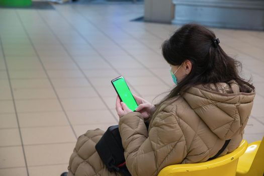 a young woman in a warm down jacket with green screen smartphone sits on an armchair in the waiting room in the subway watches the train and use social media, . High quality photo