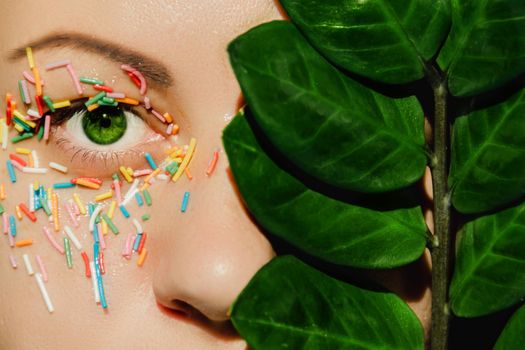 An open, green woman's eye with a sweet, multicolored sprinkle on the eyelid and a plant with large, green leaves