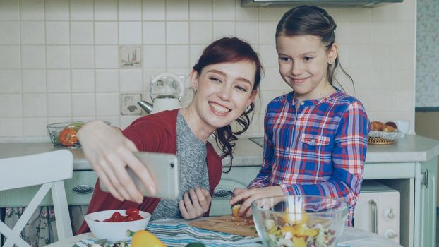Smiling mother making selfie portrait together with young cute daughter cooking breakfast at home in kitchen. Family, cook, and people concept