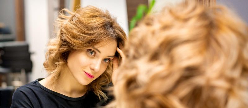 The beautiful young caucasian woman looks at her reflection in the mirror checking hairstyle and makeup in a beauty salon