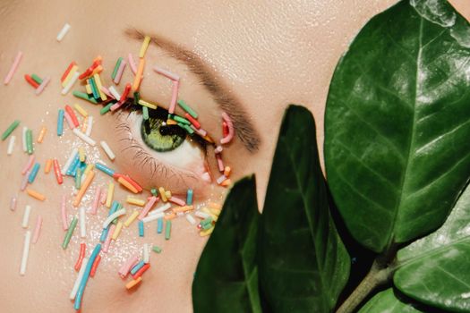 An open, green woman's eye with a sweet, multicolored sprinkle on the eyelid and a plant with large, green leaves
