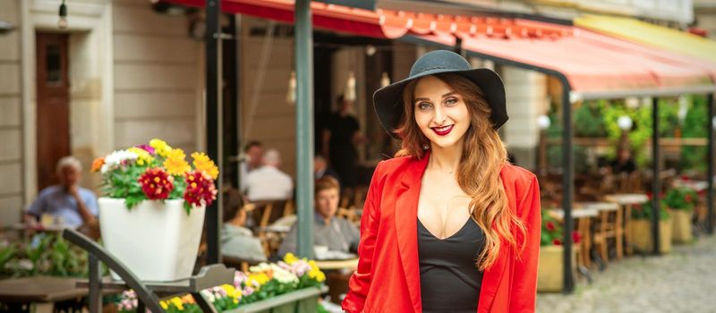 Smiling portrait of fashion young caucasian woman is posing in black hat and red jacket in the city street