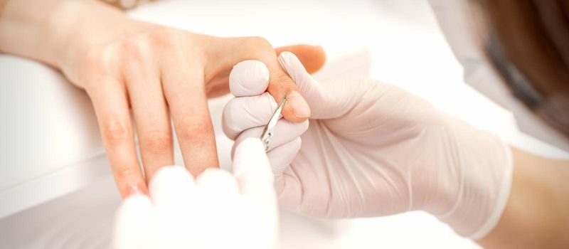 Close up of manicure master with manicure scissors removes cuticles of female nails at beauty salon