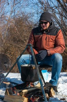 Male caucasian is sitting by campfire, pot of soot over bonfire hanging on tripod, winter outdoor cooking at campsite, lifestyle, vertikal image