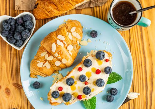 classic american breakfast. Fresh french croissant with fresh berries. blueberry and strawberry on cream cheese. Croissant and a cup with the inscription coffee on wooden background. Healthy breakfast