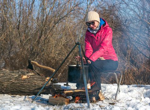 Female caucasian with sunglasses in pink jacket warms her hands by campfire, pot of soot over bonfire hanging on tripod, ax in tree trunk on backdrop, winter outdoor cooking at campsite, lifestyle