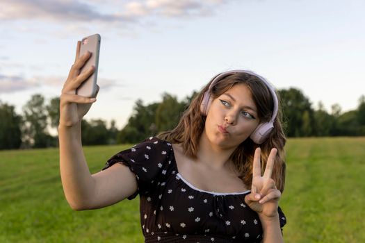 A teenage girl in a dress listens to music on her smartphone through wireless headphones and takes a selfie against a green field