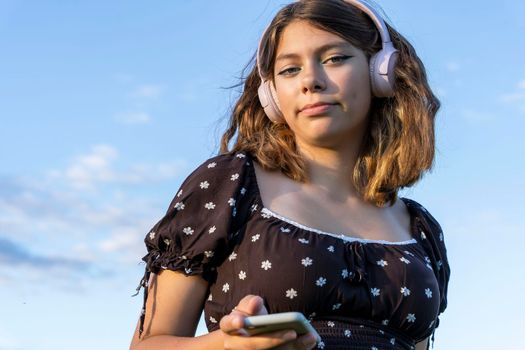 A teenage girl in a dress listens to music on her smartphone through wireless headphones against a blue sky background