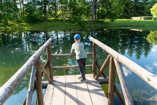 a preschooler boy is resting, leaning on the wooden bridges of the pond, walking through the nature park. A child is resting on a wooden bridge. The joys of childhood