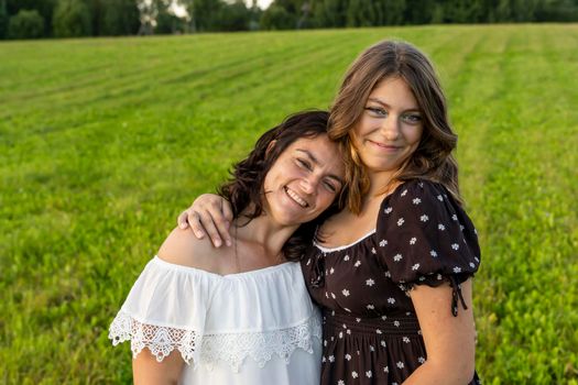 a happy mom hugs her teenage daughter in a dress against a green field. A beautiful mother and her pretty teenage daughter smile and hug. Happy Mother's Day