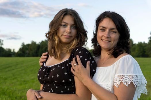 a happy mom hugs her teenage daughter in a dress against a green field. A beautiful mother and her pretty teenage daughter smile and hug. Happy Mother's Day.