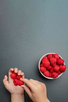 Raspberries in a saucer on a black background. The child takes raspberries from the plate. Shale stone texture. Vertical photo.