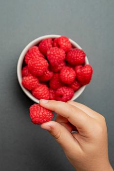 Raspberries in a saucer on a black background. The child takes raspberries from the plate. Shale stone texture. Vertical photo.