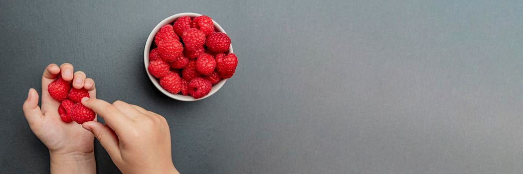 The child's hand takes fresh juicy raspberries from a small black plate. Bright red crimson close-up. Summer berry picking time. Healthy organic fruits for kids