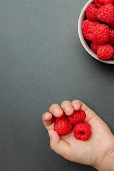 Raspberries in a saucer on a black background. The child takes raspberries from the plate. Shale stone texture. Vertical photo.