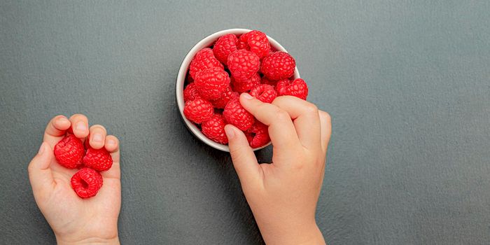 The child's hand takes fresh juicy raspberries from a small black plate. Bright red crimson close-up. Summer berry picking time. Healthy organic fruits for kids