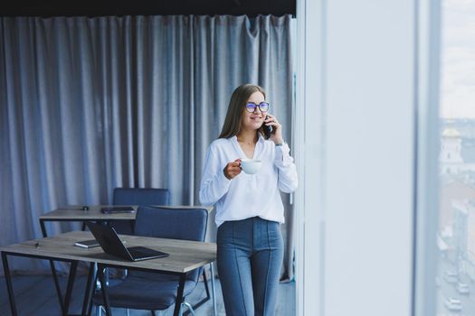 Photo of a successful young woman in a shirt smiling and working on a laptop while talking on the phone in a modern office with large windows. Remote work with coffee
