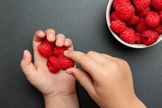 Raspberries in a saucer on a black background. The child takes raspberries from the plate and puts them in his hand. Copy space, banner on the theme of food.
