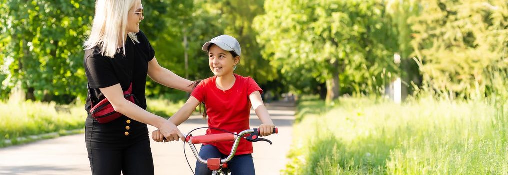 Beautiful and happy young mother teaching her daughter to ride a bicycle. Both smiling, summer park in background