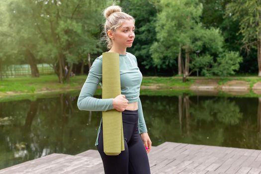 Beautiful blonde woman, holding a green gym mat, standing in the park in summer by the pond. Copy space