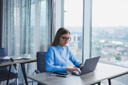 Young beautiful business woman in a blue shirt sitting in the office with a laptop and doing business