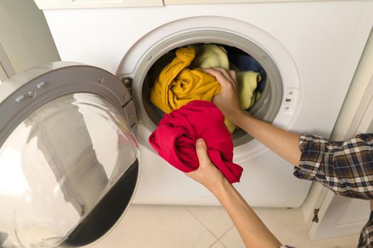 A girl folds bright clothes into a new washing machine in the bathroom at home, a woman washes things and spin them out with detergent in the laundry close-up, top view.