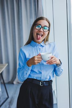Image of a happy young woman in a classic shirt and glasses smiling and drinking coffee while standing by the window in a modern office with large windows. Remote work