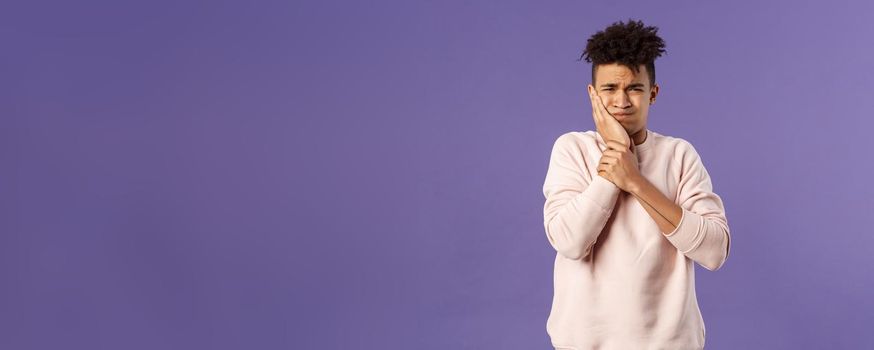 Portrait of troubled young man feeling painful ache in tooth, have toothache or decay, touching swallen cheek grimacing uneasy, need see dentist, waiting in line for appointment, purple background.