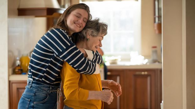 A young girl happily hugs her grandmother in a cozy house against the backdrop of the kitchen, her daughter has a good time with her mother, lovingly cares for and supports an elderly relative.