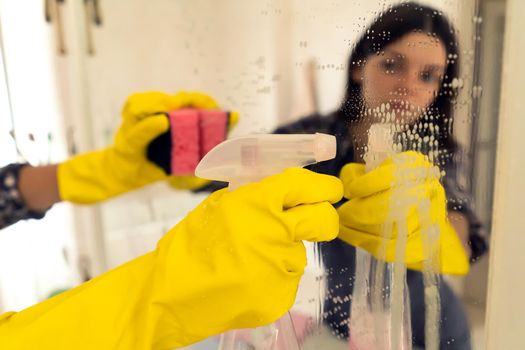 A young girl is cleaning the bathroom, applying detergent with a spray and washing the mirror with a sponge in yellow gloves on her hands. Woman taking care of the cleanliness of her home.