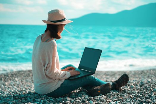 A young girl in a light hat and casual sweater lies on the beach by the sea with a laptop on a sunny day, works, studies, buys tickets during trip, a woman rests on vacation and types on the keyboard.