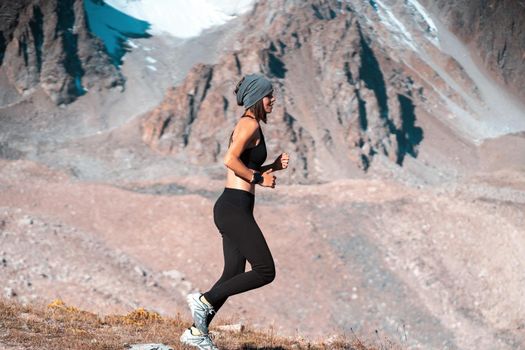 A young athletic girl jogging in a scenic area among the snow-capped mountains at sunset. A runner in leggings and a top is warming up, exercising, and involved in trail running.