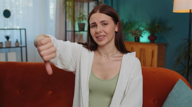 Dislike. Upset girl showing thumb down sign gesture, expressing discontent, disapproval, dissatisfied bad work at modern home apartment indoors. Displeased young woman in living room sitting on sofa