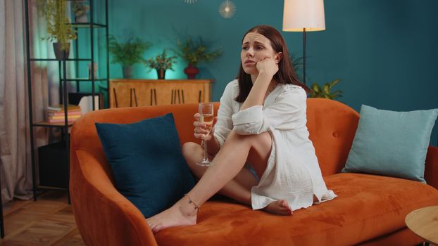 Sad young woman looks pensive of unrequited love, suffers from unfair situation, drinking champagne. Problem, break up, depressed feeling bad annoyed, burnout, bankruptcy. Girl sitting at home room