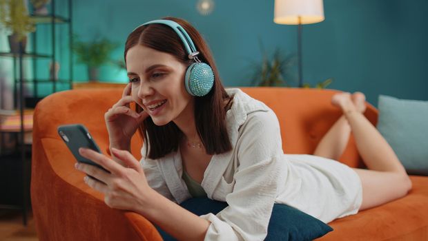 Young woman in wireless headphones relaxing lying on sofa at home choosing listening favorite energetic disco rock n roll dancing music in smartphone. People weekend activities. Happy overjoyed girl