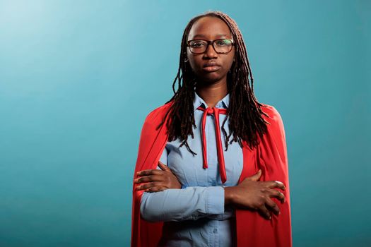 Brave and proud superhero woman wearing mighty hero cloak while standing with arms crossed on blue background. Beautiful and confident african american justice defender posing for camera. Studio shot.