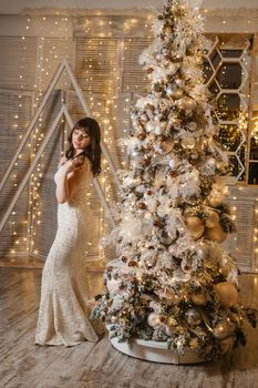 A brunette woman in a beautiful light tight dress next to a Christmas tree. The magical atmosphere of Christmas Eve on December 25. Decoration for the holiday in a light style with lots of lights and light bulbs.