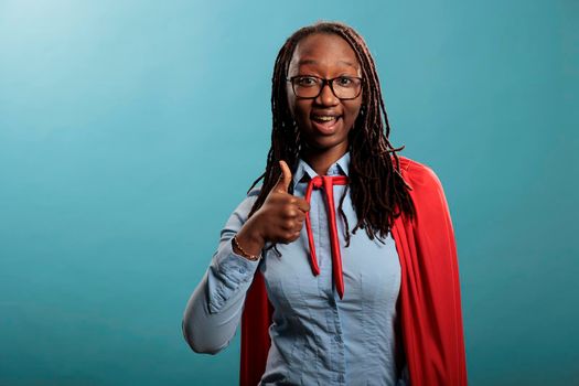 Brave and proud superhero woman wearing cloak while giving thumbs up gesture sign on blue background. Strong and confident african american justice defender expressing ok symbol at camera. Studio shot