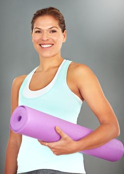 On her way to yoga class. Portrait of a young woman holding a yoga mat while standing against a gray background