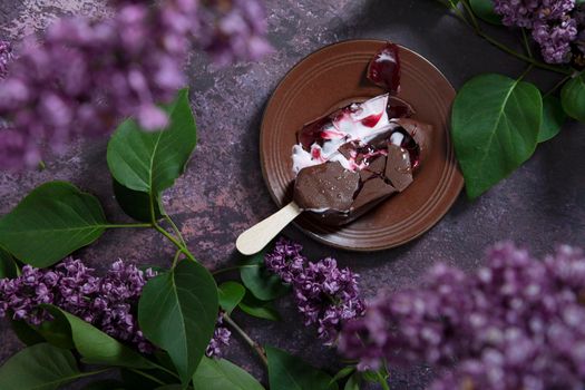 melted raspberry ice cream in chocolate on a wooden stick on a dark burgundy background with lilac flowers, a sweet dessert on a hot spring day, flat lay food. High quality photo