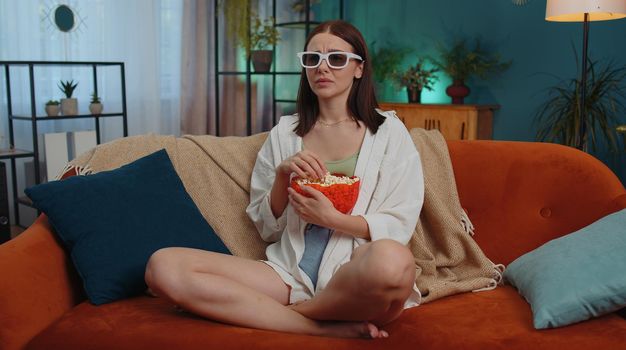 Excited adult girl sitting on sofa eating popcorn and watching interesting tv serial, sport game, film, online social media movie content online at home. Young woman enjoying domestic entertainment