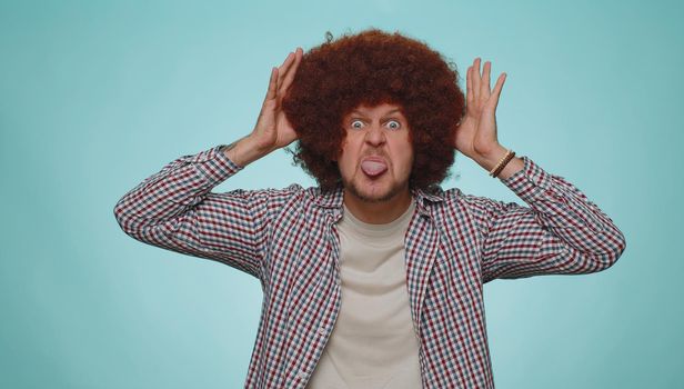 Cheerful funny man with lush hairstyle showing tongue making faces at camera, fooling around, joking, aping with silly face, teasing. Young adult guy boy isolated alone on blue studio background