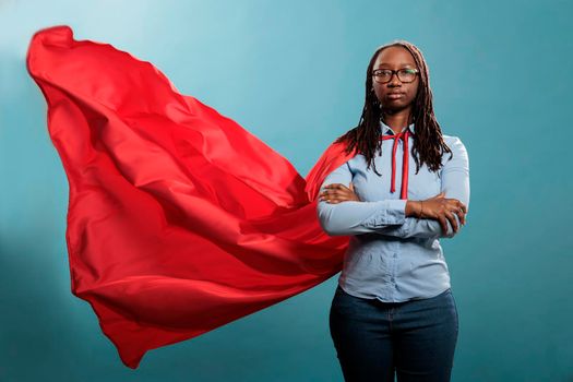 Confident and brave young superhero woman standing with arms crossed while wearing red mighty cape on blue background. Portrait of justice defender posing tough and strong while looking at camera.